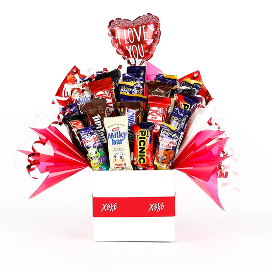 Chocolate bouquet with various chocolate bars & a "I Love You" balloon in a box labeled 'XOXO' on a white background