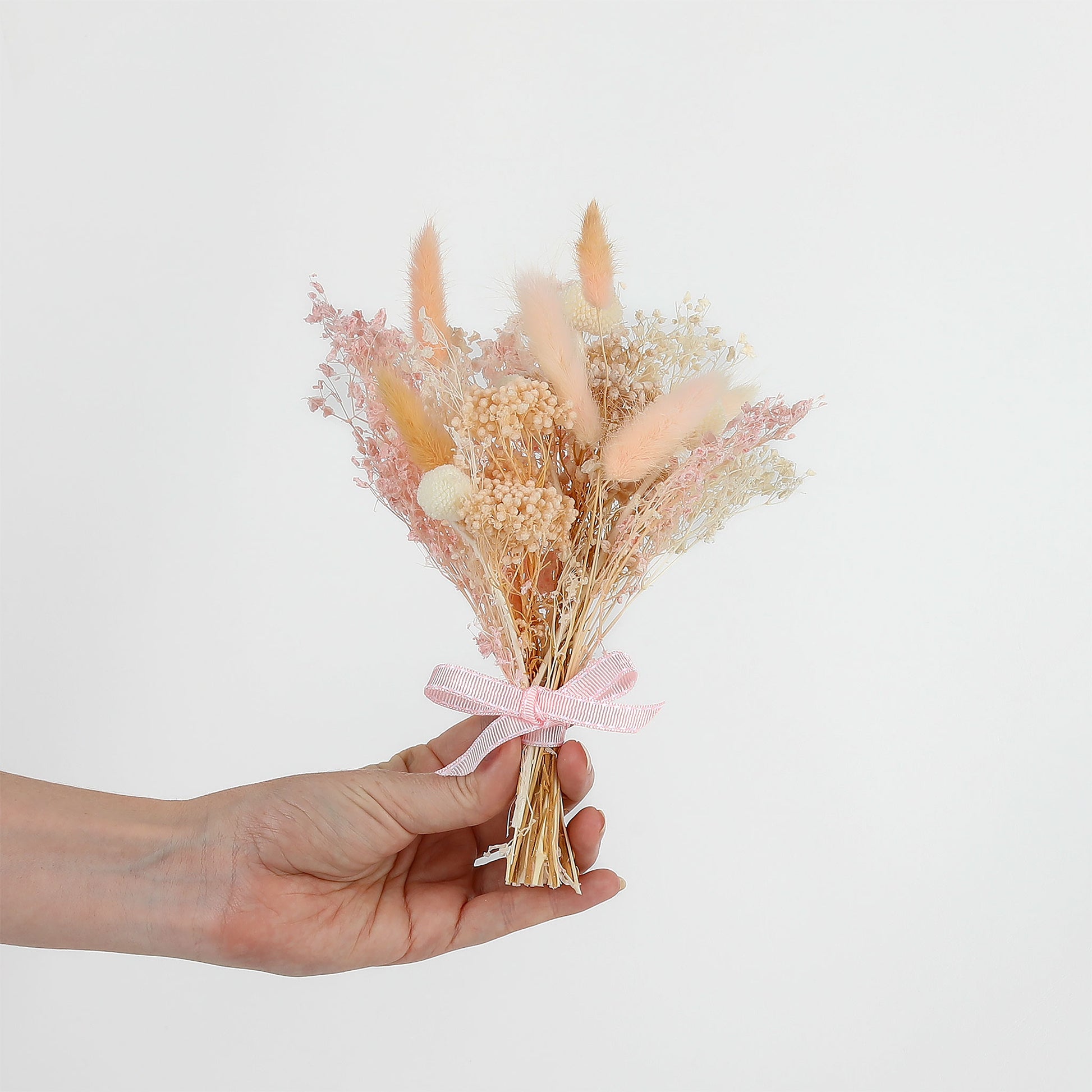 Hand holding a small bouquet of dried flowers with a pink ribbon against a light background
