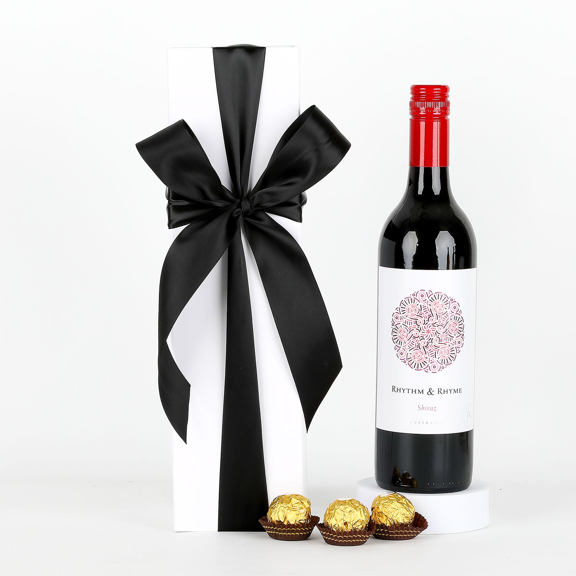 Bottle of red wine with a black ribbon, white box, and gold chocolates on a white background