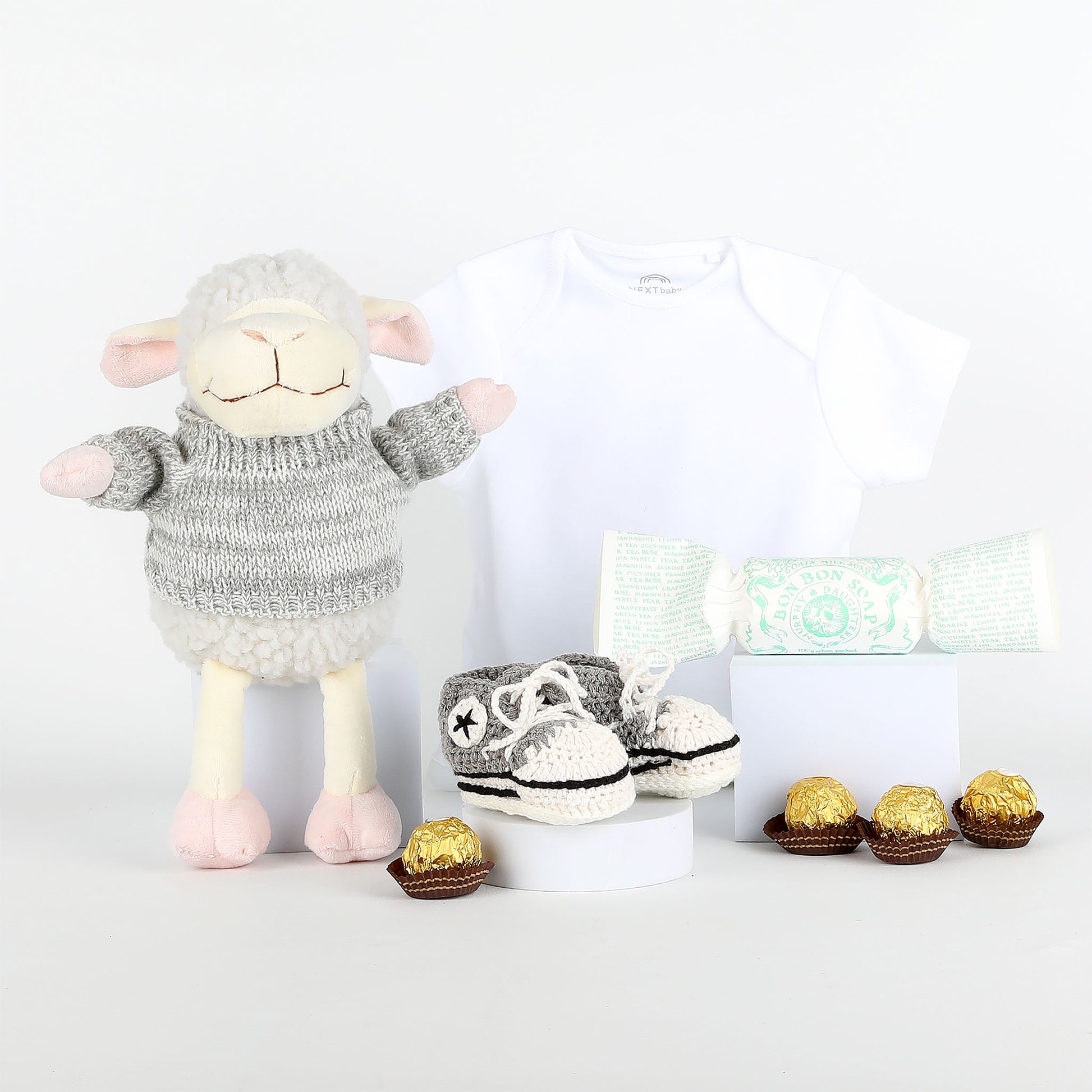 Plush lamb toy wearing a sweater with baby shoes and chocolates on a white background